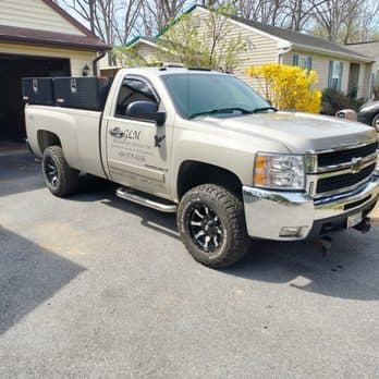 A white GMC pickup truck with "GLM Mechanical Services, Inc." branding parked on a residential street, featuring a black utility bed and black wheels, with houses and trees in the background.